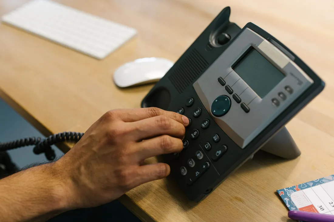 Hand wählt eine Nummer auf einem schwarzen Tischtelefon neben Tastatur und Maus